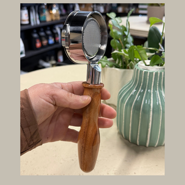 Hand holding a wooden-handled coffee grinder with a metal top, blurred indoor setting with plants and bottles.